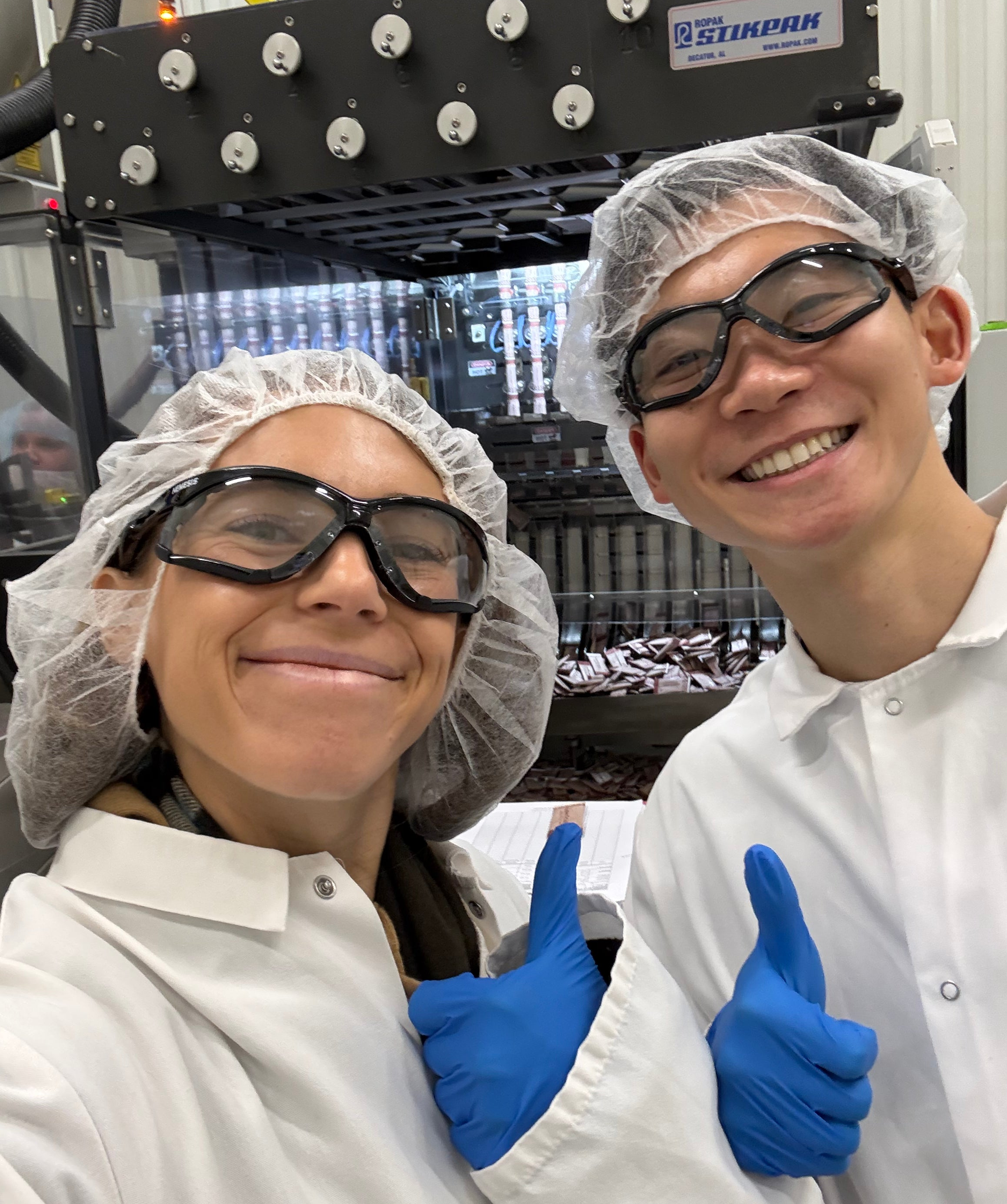 Two coffee production team members wearing lab coats, hair nets, safety goggles, and gloves giving thumbs up inside a clean food manufacturing facility, highlighting quality control and clean ingredients in coffee production.