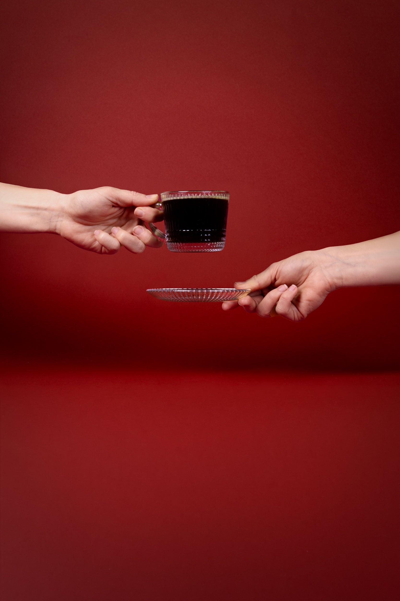 Minimalist image of hands exchanging a glass cup of black coffee on a red backdrop.