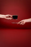 Minimalist image of hands exchanging a glass cup of black coffee on a red backdrop.