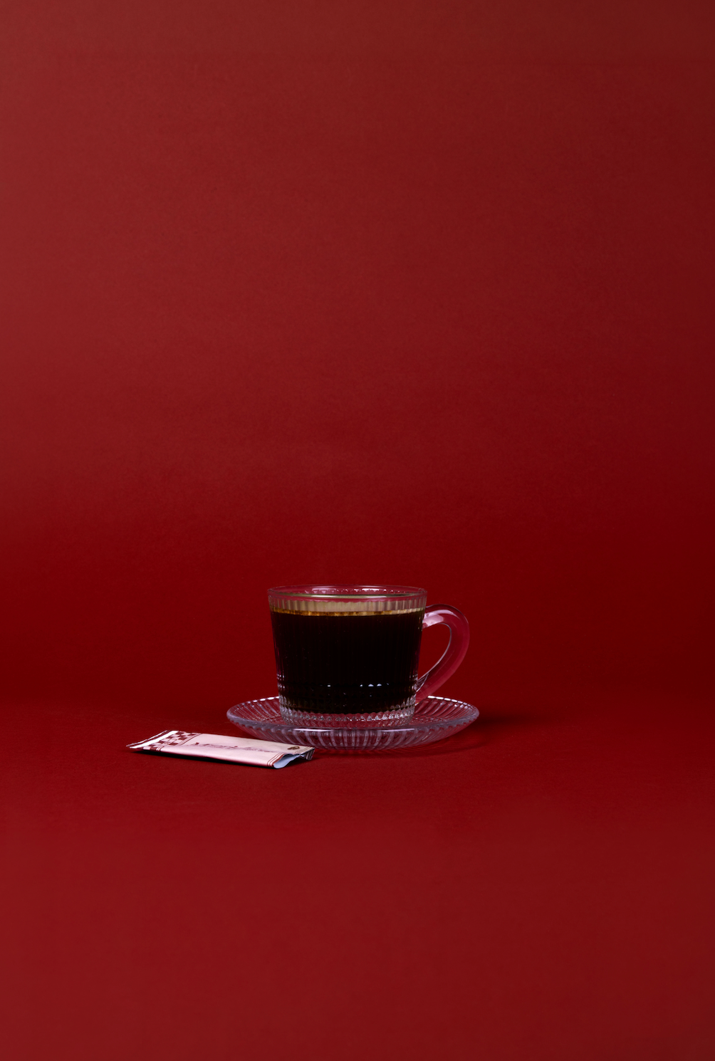 Glass cup of black coffee on a clear saucer against a bold red background, minimalist morning coffee setup highlighting simple ingredients and portable coffee routine.