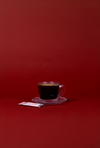 Glass cup of black coffee on a clear saucer against a bold red background, minimalist morning coffee setup highlighting simple ingredients and portable coffee routine.