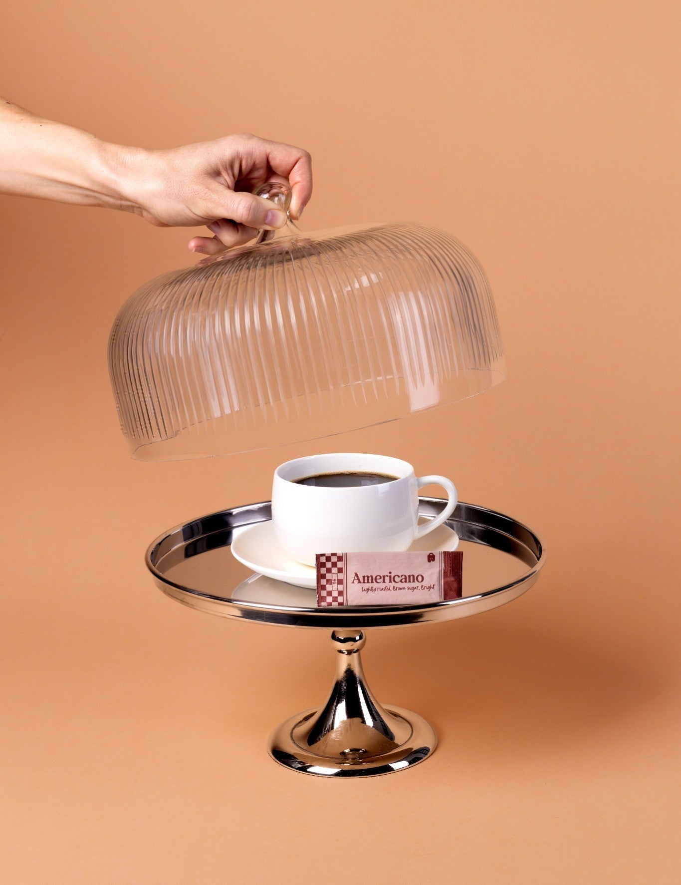 Hand holding a glass cloche over a cup of coffee on a cake stand with a brown background