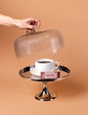 Hand holding a glass cloche over a cup of coffee on a cake stand with a brown background
