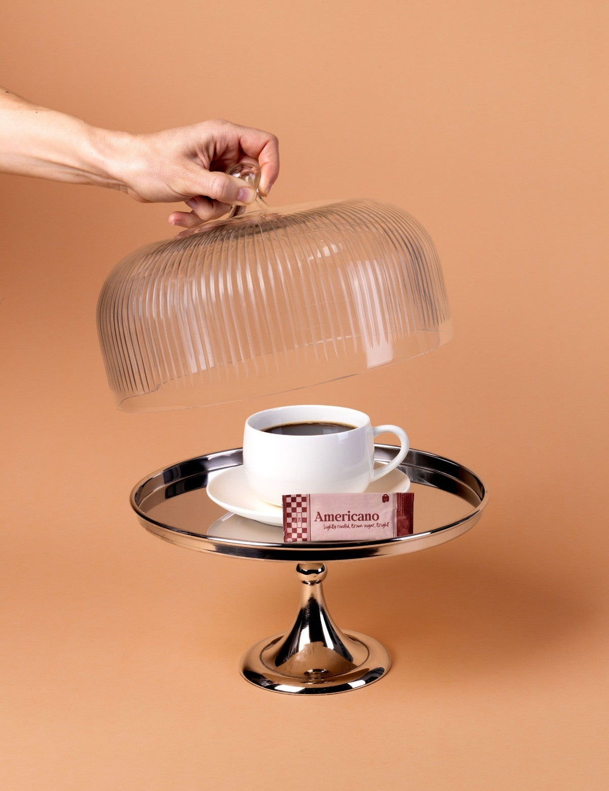 Hand holding a glass cloche over a cup of coffee on a cake stand with a brown background